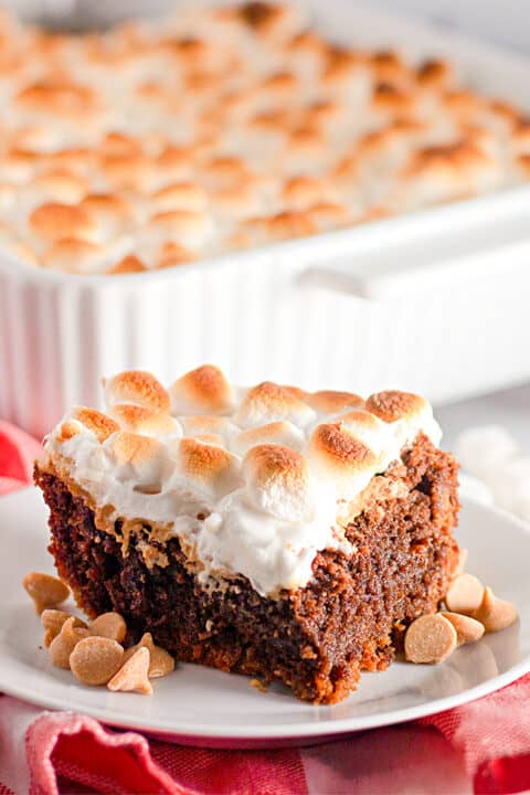A square piece of Peanut Butter S'mores Brownie, topped with toasted marshmallows, sits on a white plate with butterscotch chips scattered beside it. A baking dish of brownies is in the background.