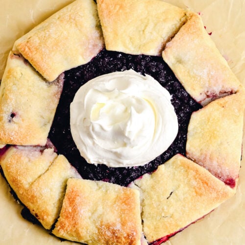 A rustic tart with a golden, folded crust is filled with dark berries in this Blueberry Galette, topped with a swirl of whipped cream and set on parchment paper.