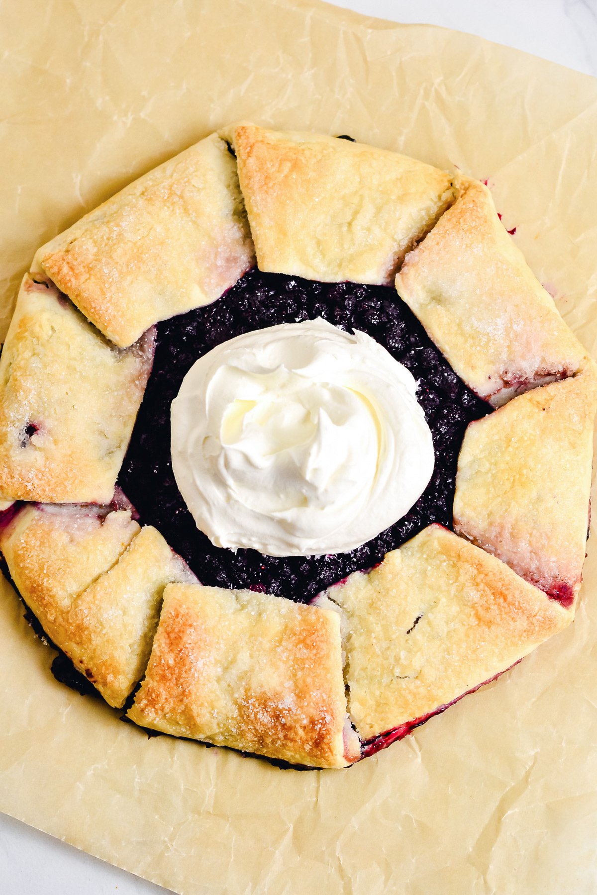 A rustic tart with a golden, folded crust is filled with dark berries in this Blueberry Galette, topped with a swirl of whipped cream and set on parchment paper.