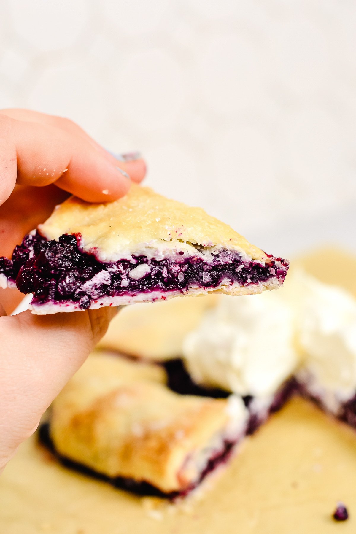 A hand holds a slice of blueberry galette with a golden crust; more rustic pie and a dollop of whipped cream or ice cream are on a plate in the background.