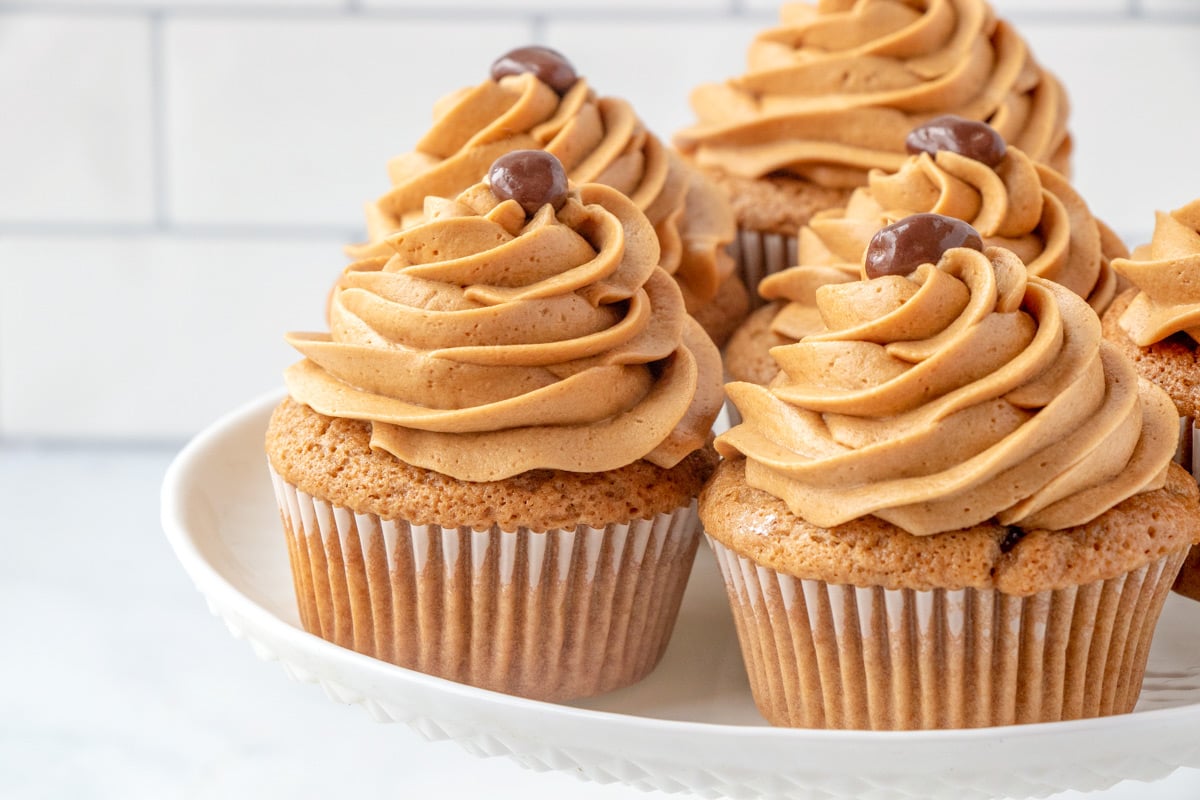 Four cupcakes with swirled coffee buttercream frosting and a small round chocolate on top, displayed on a white plate with a tiled background.