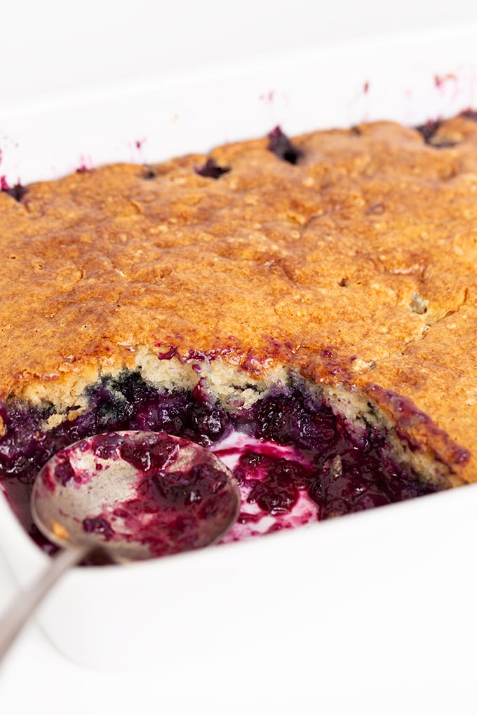 A close-up of an Easy Blueberry Cobbler in a white dish, with a serving spoon and one portion already removed.