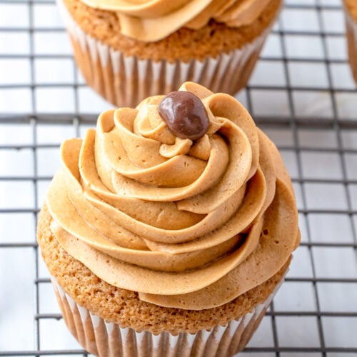 A cupcake with coffee frosting, topped with a small round chocolate, sits on a wire cooling rack.