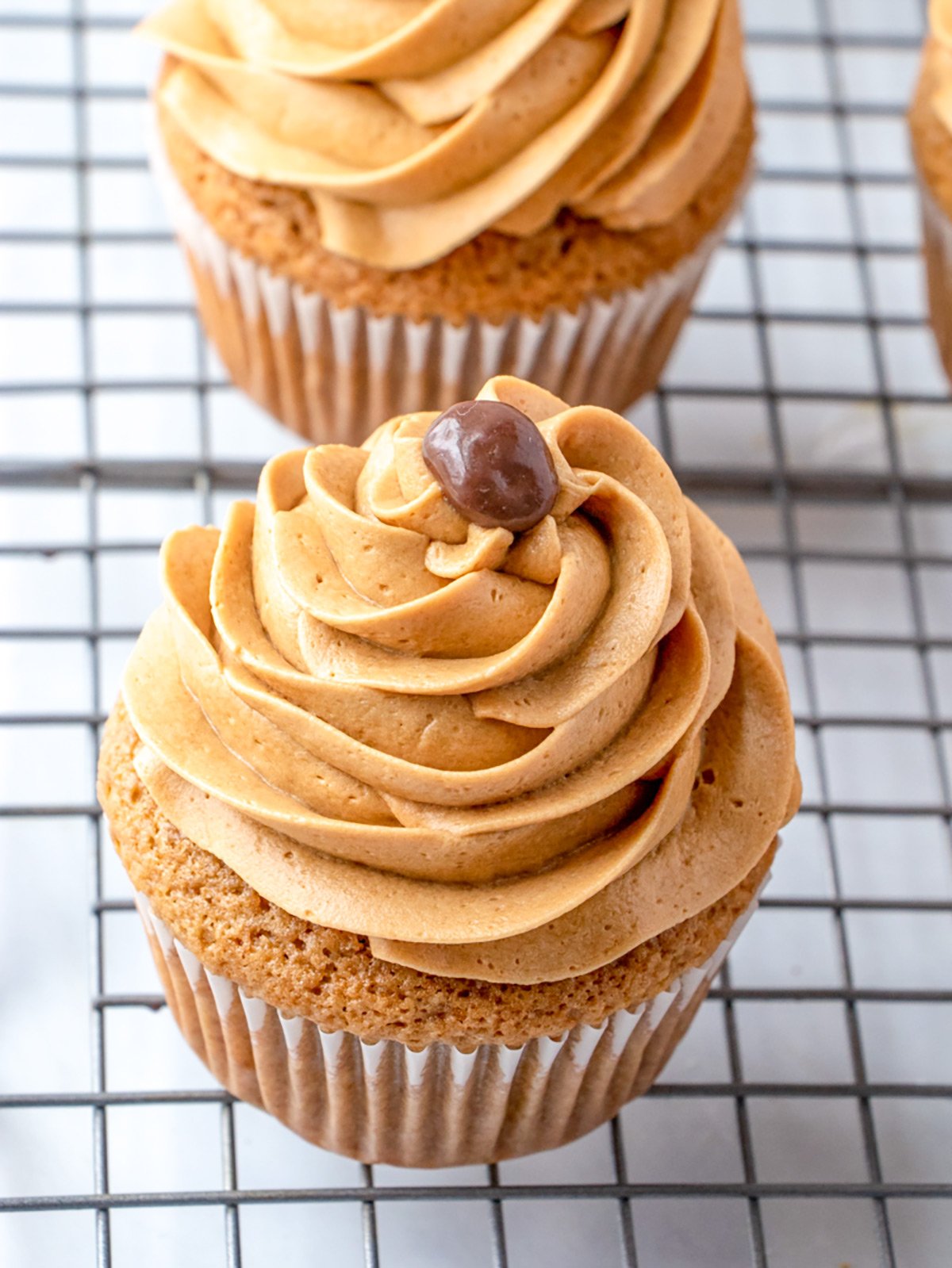 A cupcake with coffee frosting, topped with a small round chocolate, sits on a wire cooling rack.