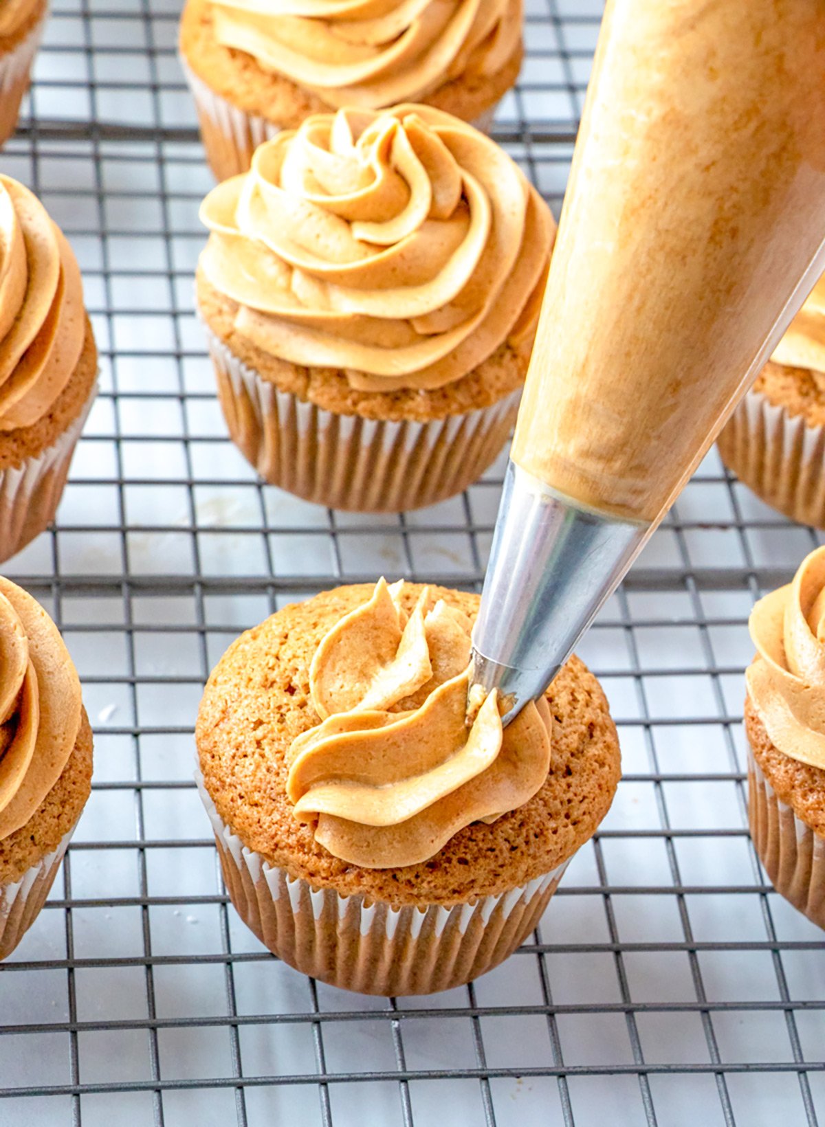 A piping bag is being used to add swirls of rich coffee frosting onto a cupcake, with several beautifully frosted cupcakes on a wire cooling rack in the background.