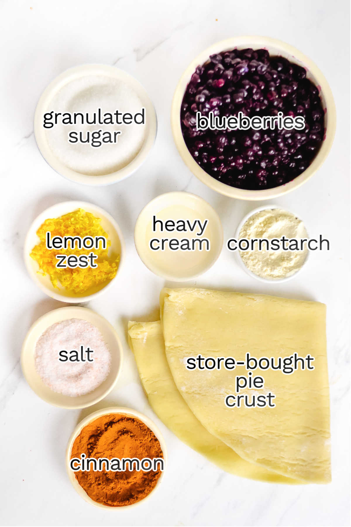 Bowls containing ingredients for a Blueberry Galette—granulated sugar, blueberries, lemon zest, heavy cream, cornstarch, salt, cinnamon, and sheets of store-bought pie crust—are arranged on a white surface for this easy blueberry dessert.