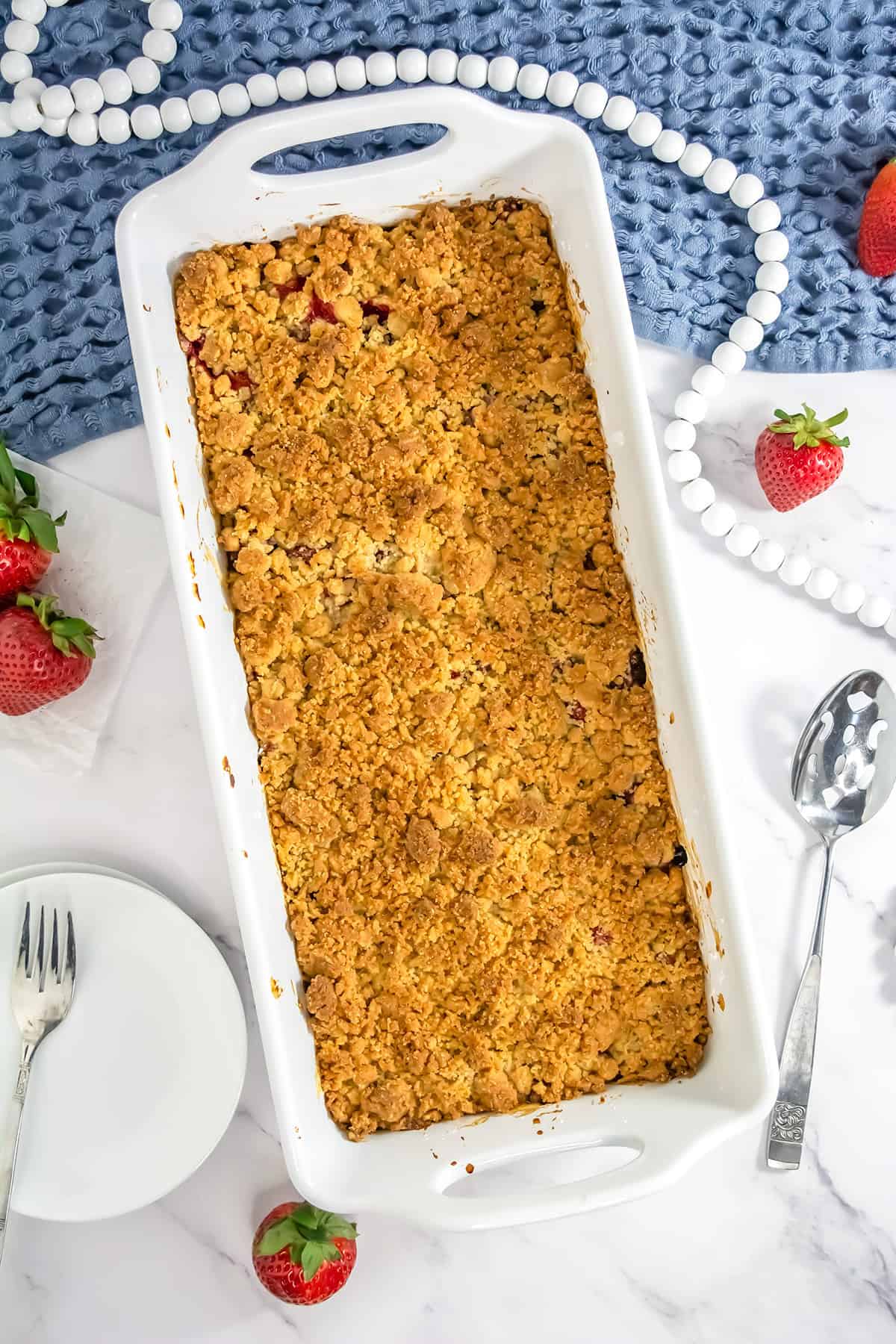 A rectangular white baking dish filled with golden Strawberry Rhubarb Crisp sits on a marble surface, surrounded by strawberries, a serving spoon, plates, and forks.