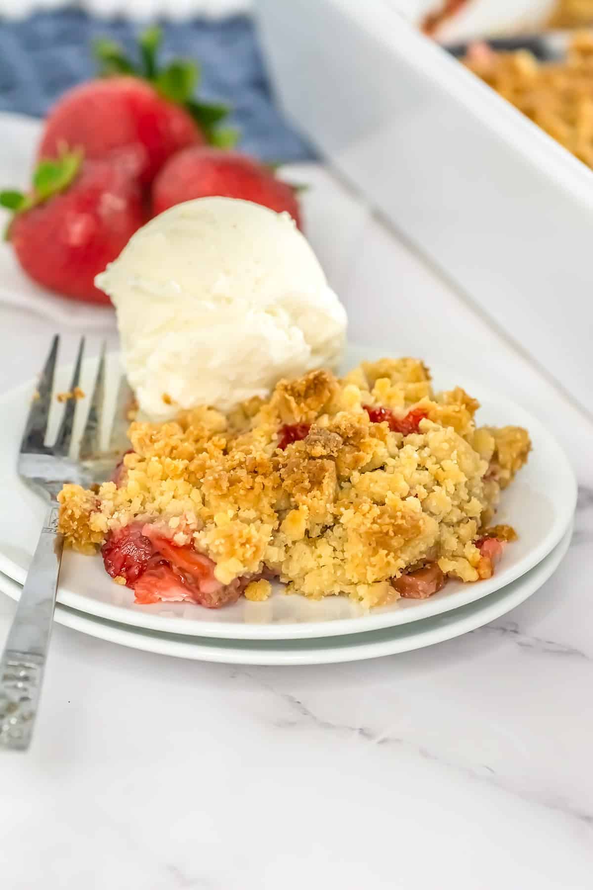A plate of Strawberry Rhubarb Crisp with a scoop of vanilla ice cream and a fork, with fresh strawberries in the background.
