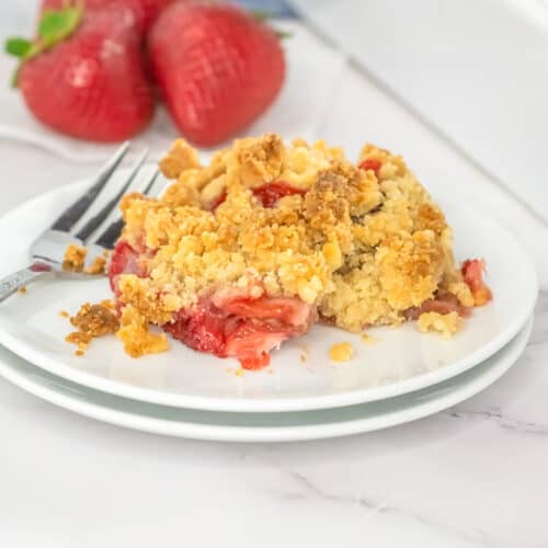 A serving of strawberry rhubarb crisp with a crumbly topping on a white plate, accompanied by a fork, with fresh strawberries in the background.