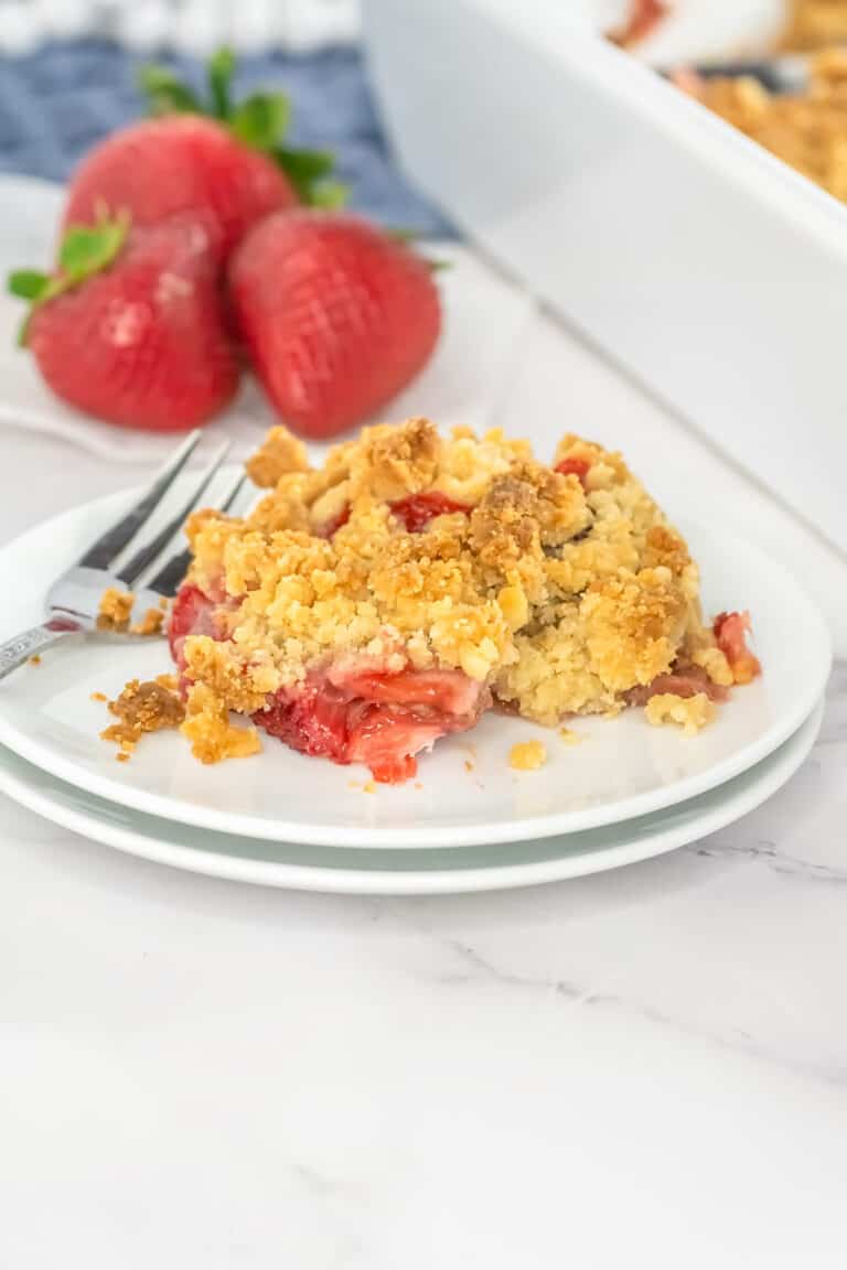 A serving of strawberry rhubarb crisp with a crumbly topping on a white plate, accompanied by a fork, with fresh strawberries in the background.
