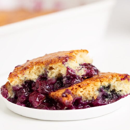 A serving of Easy Blueberry Cobbler with a golden-brown crust and juicy blueberries sits on a white plate.