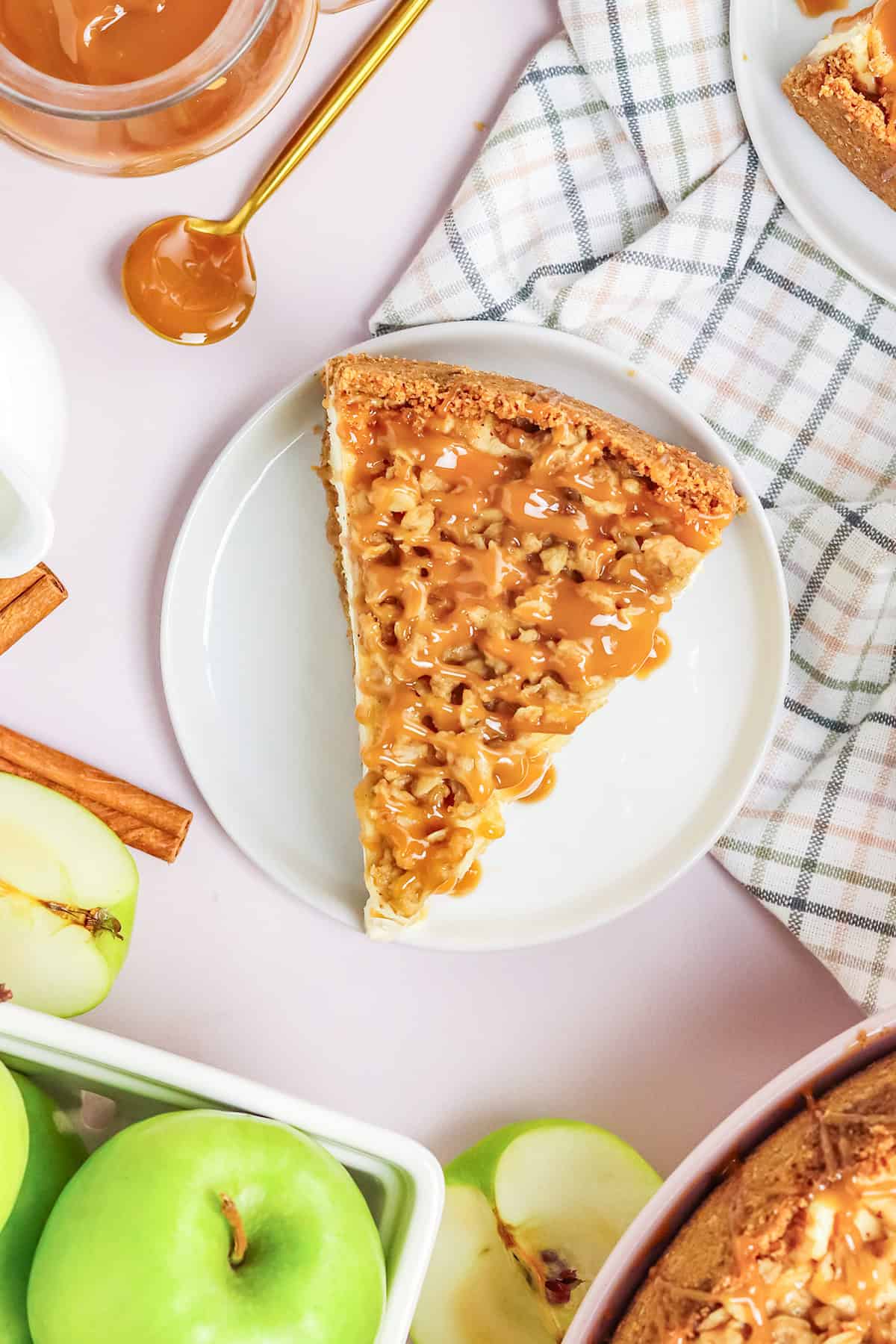 A slice of caramel apple cheesecake on a white plate, surrounded by green apples, caramel sauce, and a checkered cloth—perfect for anyone who loves the flavors of apple crisp cheesecake.