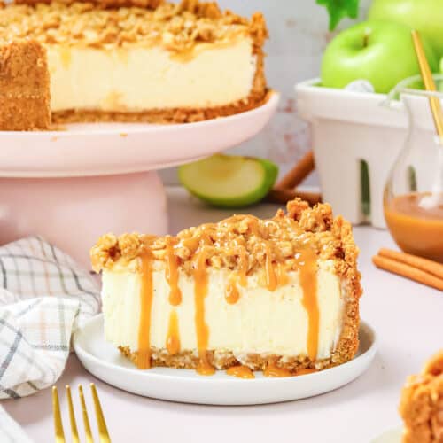 A slice of Apple Crisp Cheesecake with caramel drizzle and crumb topping sits on a plate, while the rest of the cheesecake and fresh green apples are visible in the background.