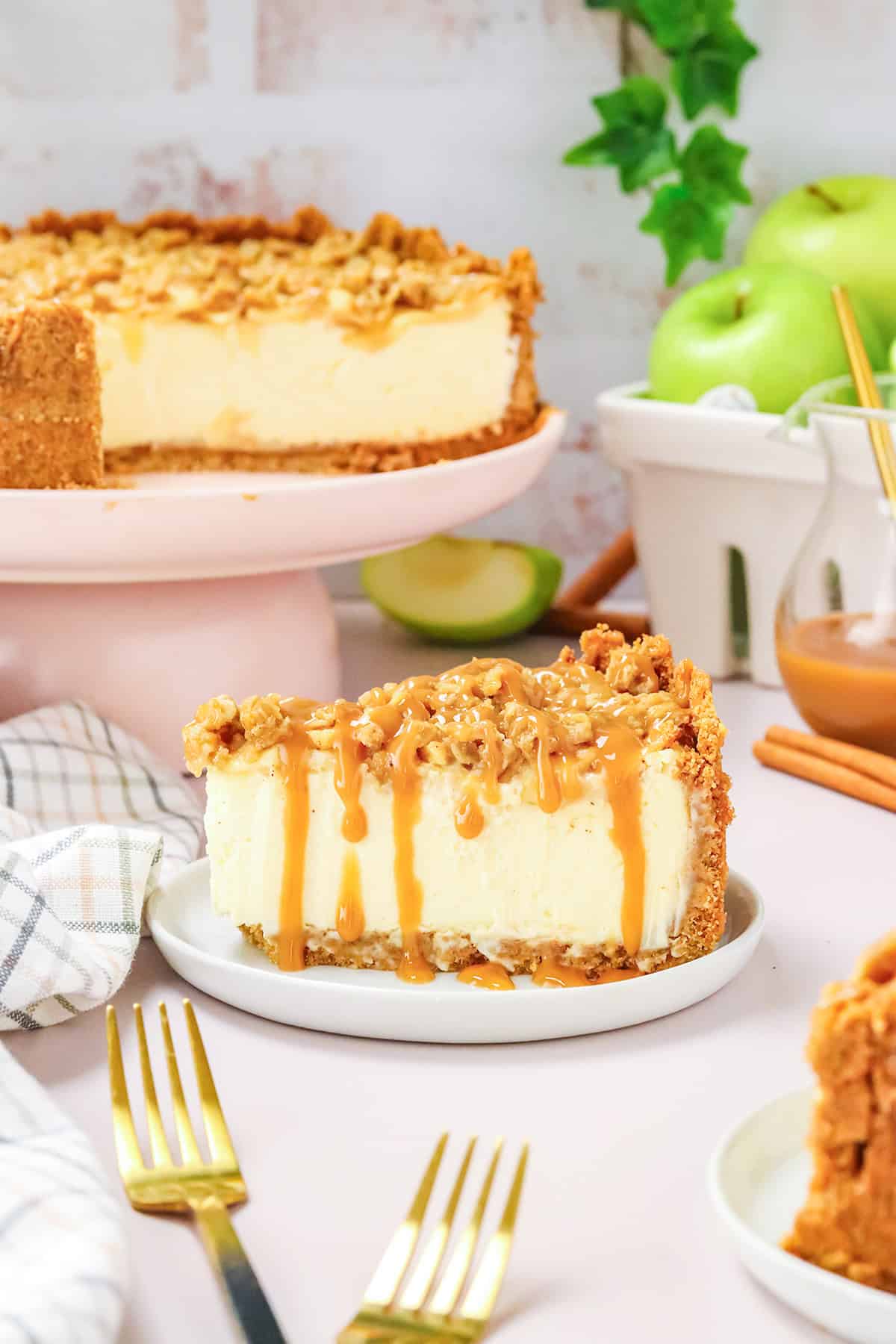 A slice of Apple Crisp Cheesecake with caramel drizzle and crumb topping sits on a plate, while the rest of the cheesecake and fresh green apples are visible in the background.