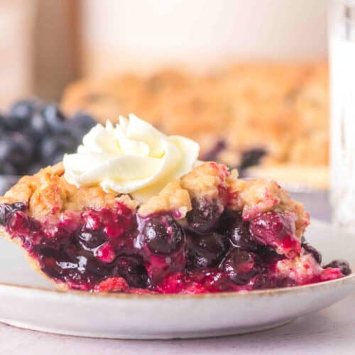 A slice of blueberry pie with a golden crust and whipped cream on top sits on a white plate, with a blurred background featuring more blueberry pie and a glass.