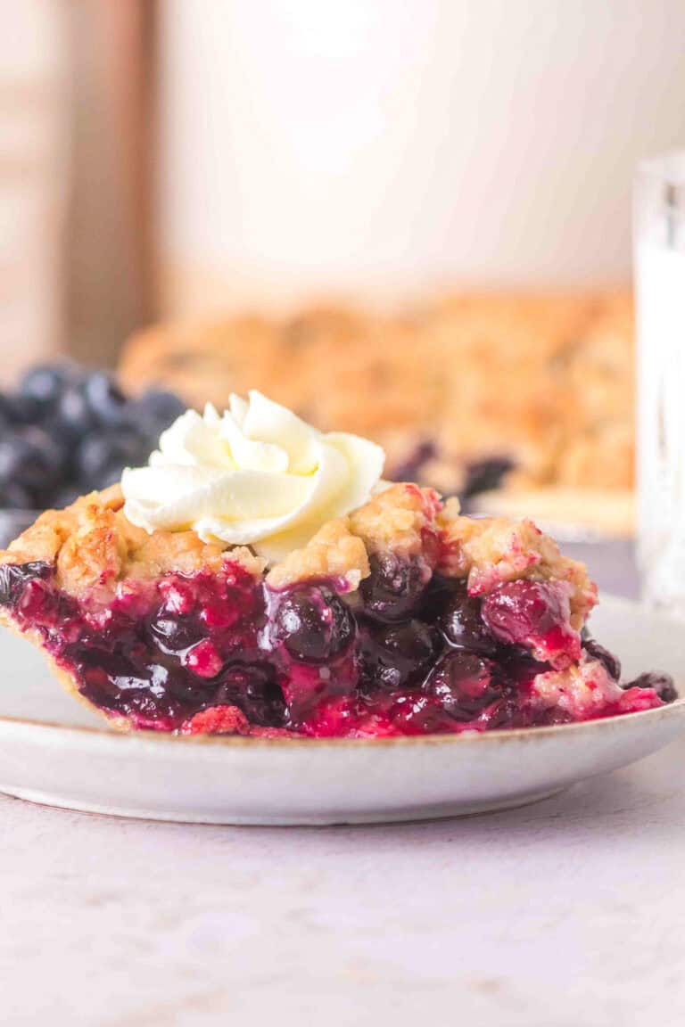 A slice of blueberry pie with a golden crust and whipped cream on top sits on a white plate, with a blurred background featuring more blueberry pie and a glass.