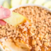 A hand holds an apple slice dipped in creamy Caramel Apple Dip with nuts, while a bowl of the decadent dessert and more fresh apple slices are arranged in the background.