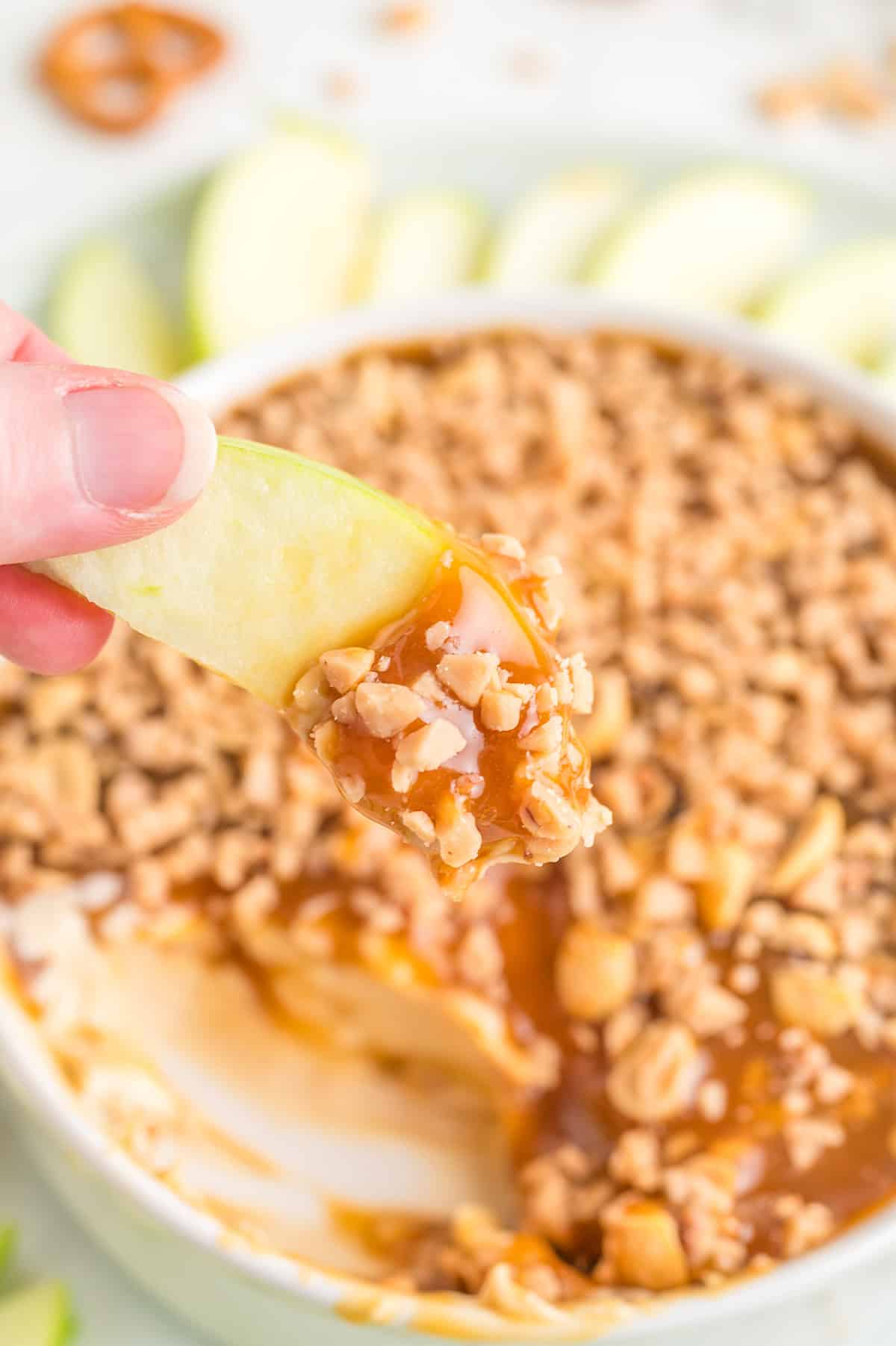 A hand holds an apple slice dipped in creamy Caramel Apple Dip with nuts, while a bowl of the decadent dessert and more fresh apple slices are arranged in the background.