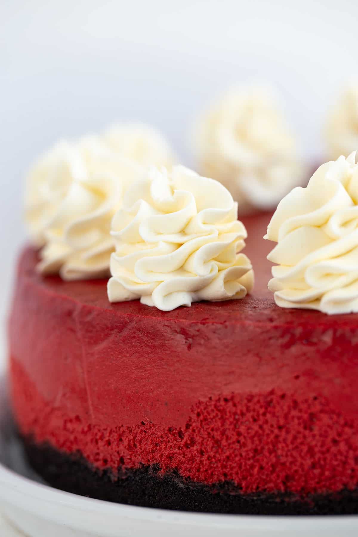 A close-up of a red velvet cake topped with large swirls of cream cheese frosting, showing its dense texture and smooth surface.
