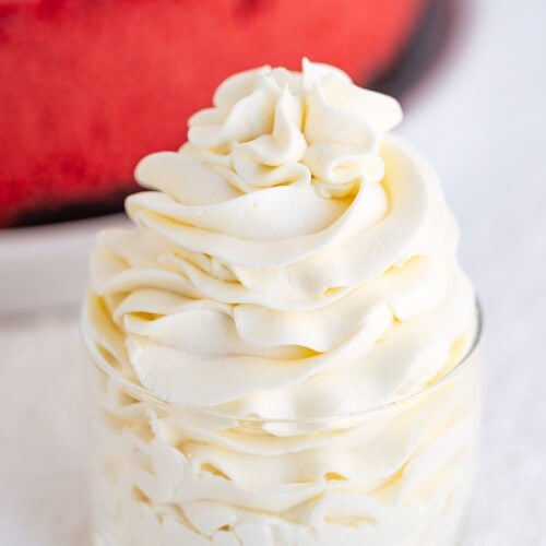 A small glass cup filled with swirled cream cheese whipped cream, with a blurred red cake in the background.