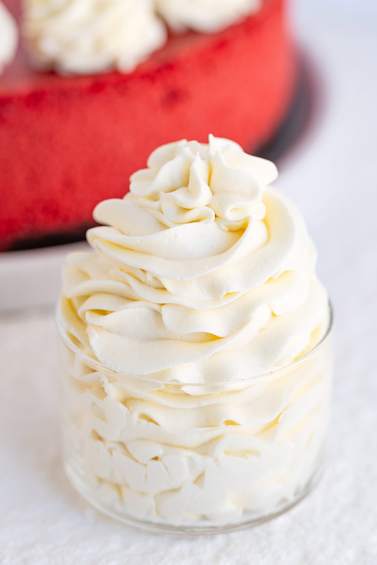 A small glass cup filled with swirled cream cheese whipped cream, with a blurred red cake in the background.