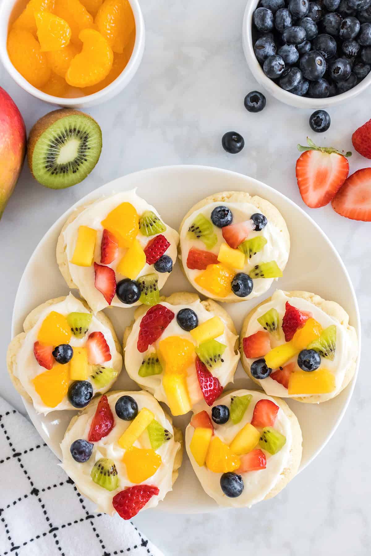 A plate of Fruit Pizza Cookies topped with cream and assorted fresh fruit, surrounded by bowls of blueberries and mandarin oranges, plus sliced strawberries, kiwi, and mango on a white surface.