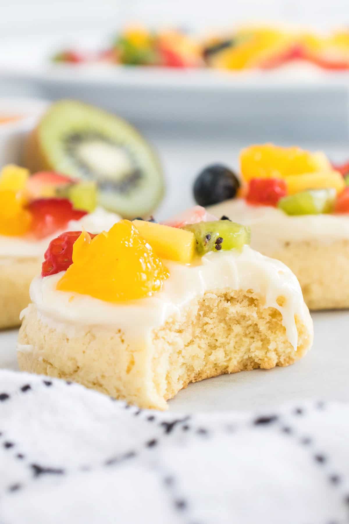 A close-up of a frosted fruit pizza cookie topped with assorted fresh fruit, with a bite taken out, resting on a white surface near a patterned cloth.