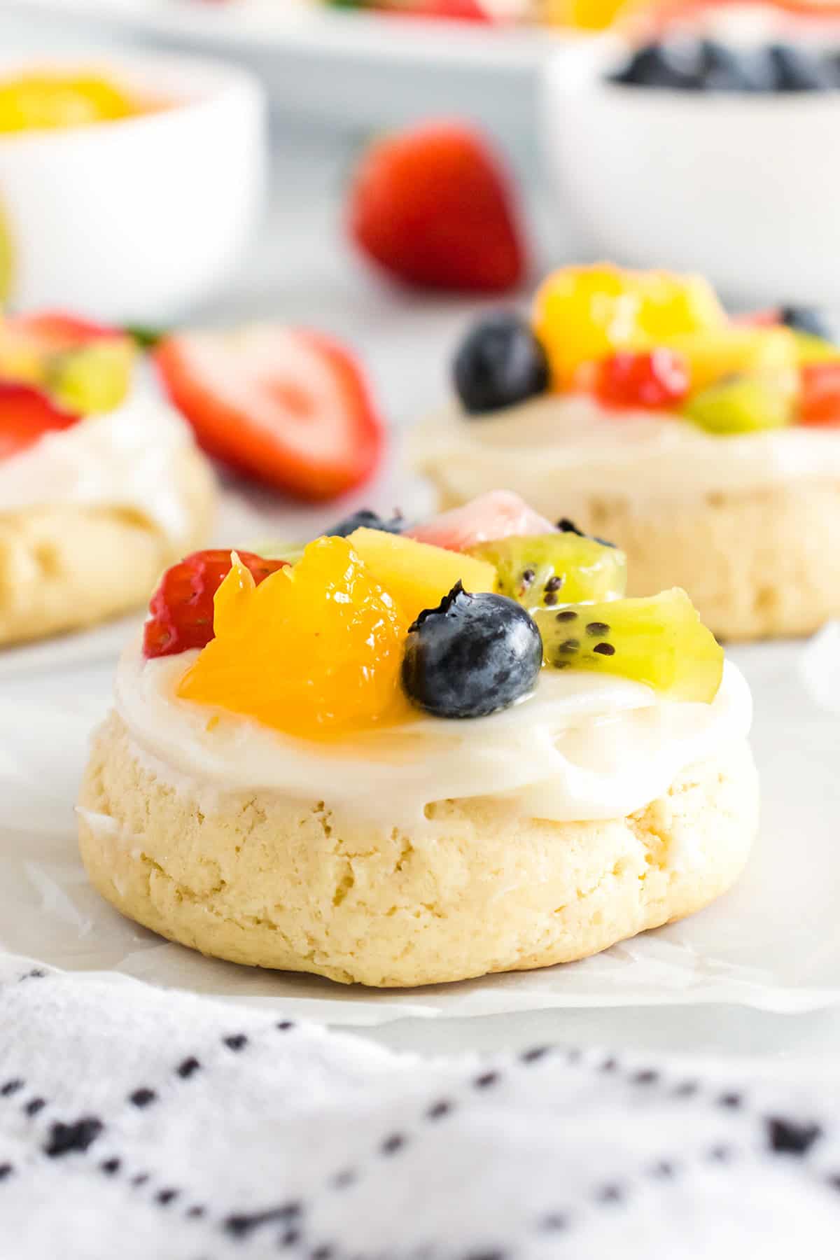 A round sugar cookie topped with white frosting and assorted fresh fruit—blueberries, kiwi, and orange segments—creates a colorful fruit pizza cookie on a white surface.