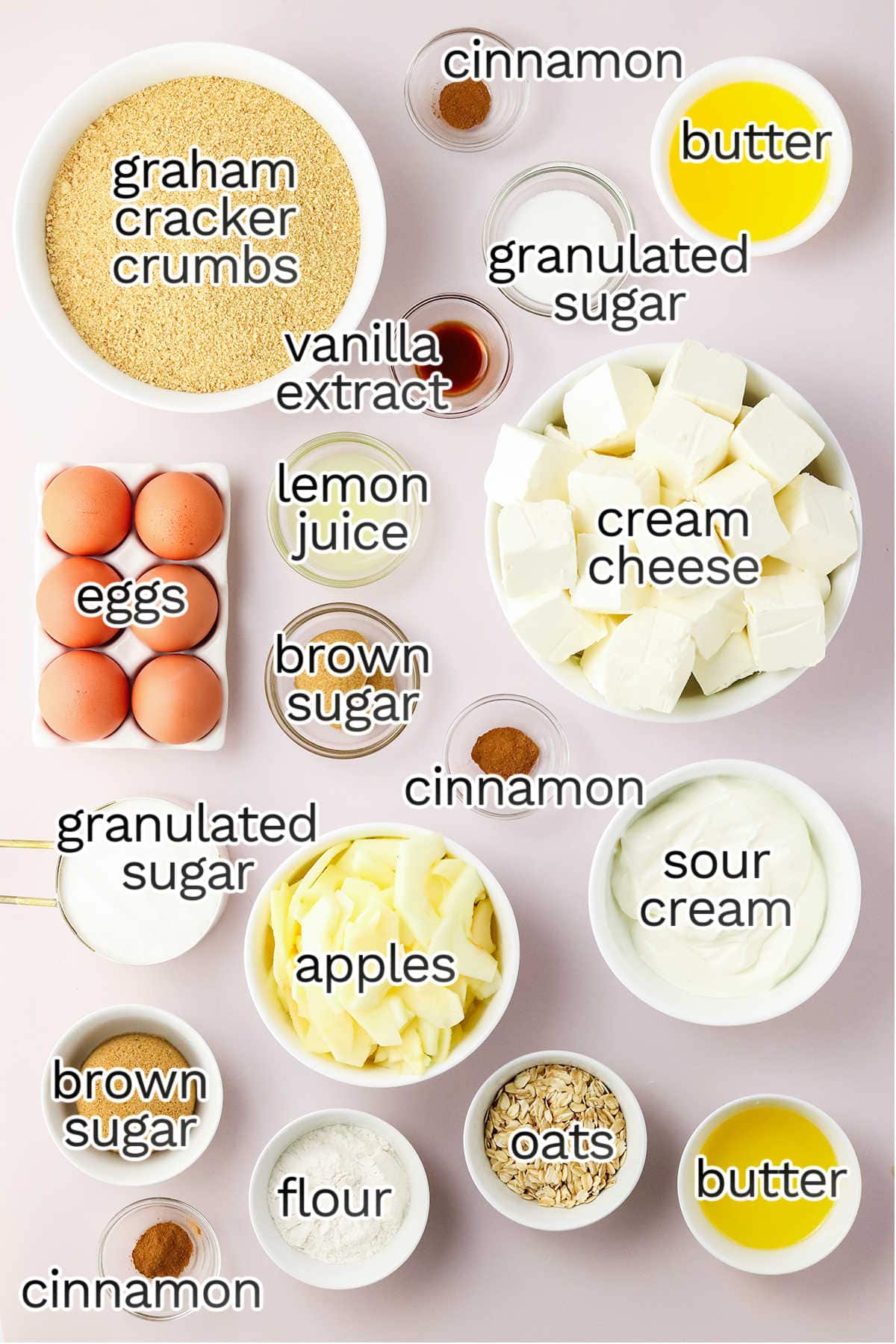 Top view of labeled baking ingredients in bowls for Apple Crisp Cheesecake, including graham cracker crumbs, cream cheese, eggs, apples, sugars, oats, flour, sour cream, butter, lemon juice, vanilla, and cinnamon.