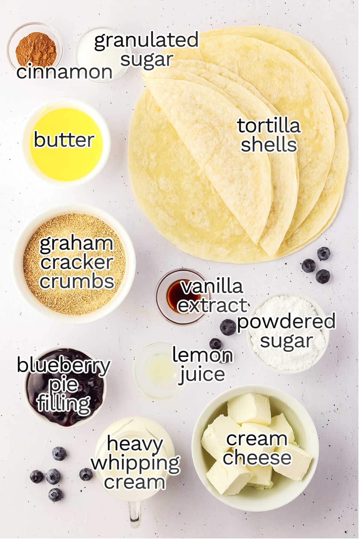 Top-down view of ingredients for a Blueberry Cheesecake Tacos dessert, featuring tortilla shells, butter, cream cheese, heavy whipping cream, blueberry pie filling, graham cracker crumbs, cinnamon, and assorted sugars.