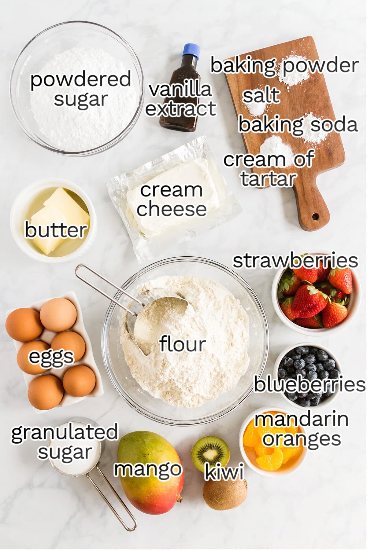 An assortment of baking ingredients and fruits labeled on a white surface, perfect for making Fruit Pizza Cookies, including flour, eggs, butter, cream cheese, sugar, vanilla, and various fresh fruits.