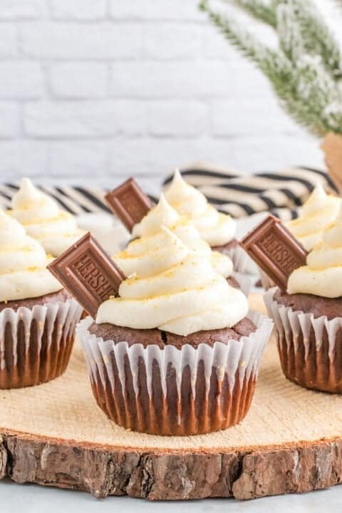 Four chocolate cupcakes with white frosting and a piece of Hershey's chocolate on top, displayed on a wooden slab against a light background—perfect for anyone searching for the ultimate S'mores cupcakes.
