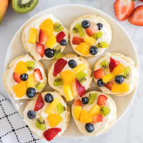 A white plate holds seven fruit pizza cookies, each topped with cream and assorted fresh fruit pieces like kiwi, strawberry, blueberry, pineapple, and orange.