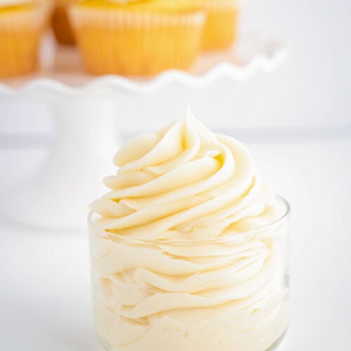 Pipine white cream cheese frosting on a glass bowl on a white surface with a cake plate behind it