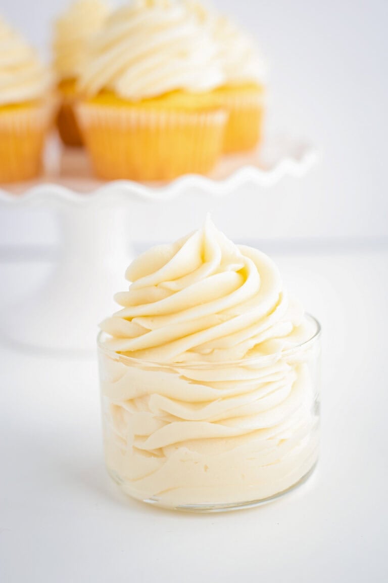 Pipine white cream cheese frosting on a glass bowl on a white surface with a cake plate behind it