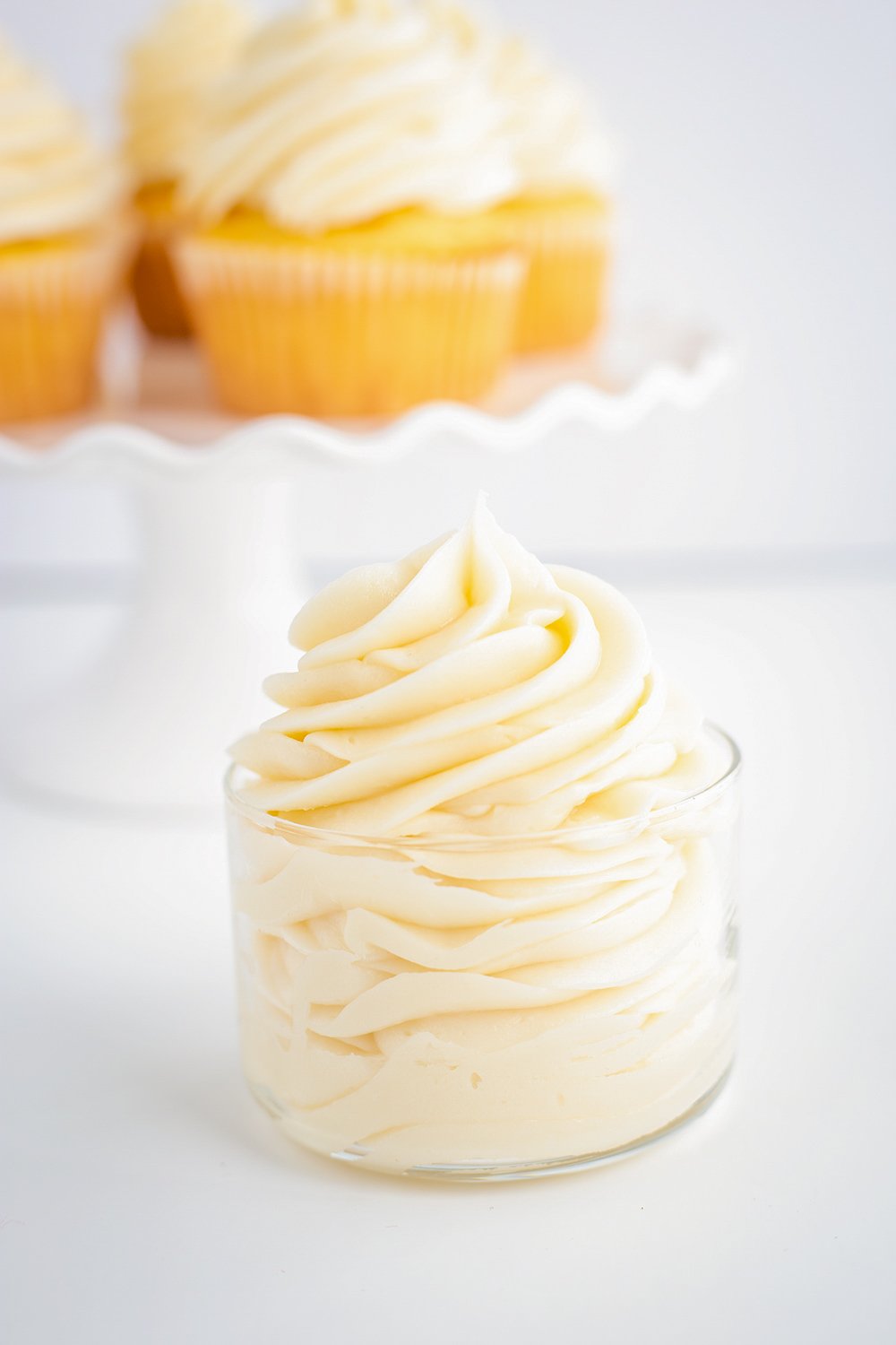 Pipine white cream cheese frosting on a glass bowl on a white surface with a cake plate behind it