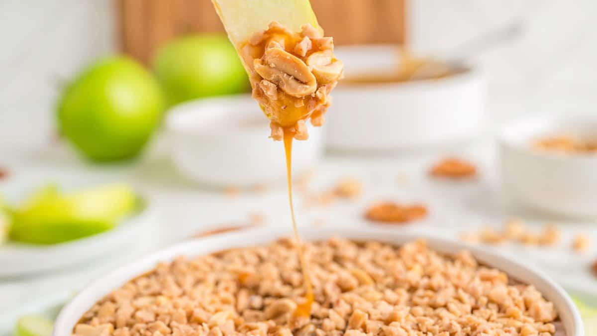 A slice of green apple dipped in caramel and chopped nuts is held above a dish filled with more chopped nuts, making it a perfect treat for autumn desserts, with apples and bowls in the background.