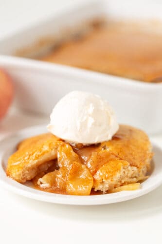 A serving of warm Apple Cobbler topped with a scoop of vanilla ice cream on a white plate, with the baking dish in the background.