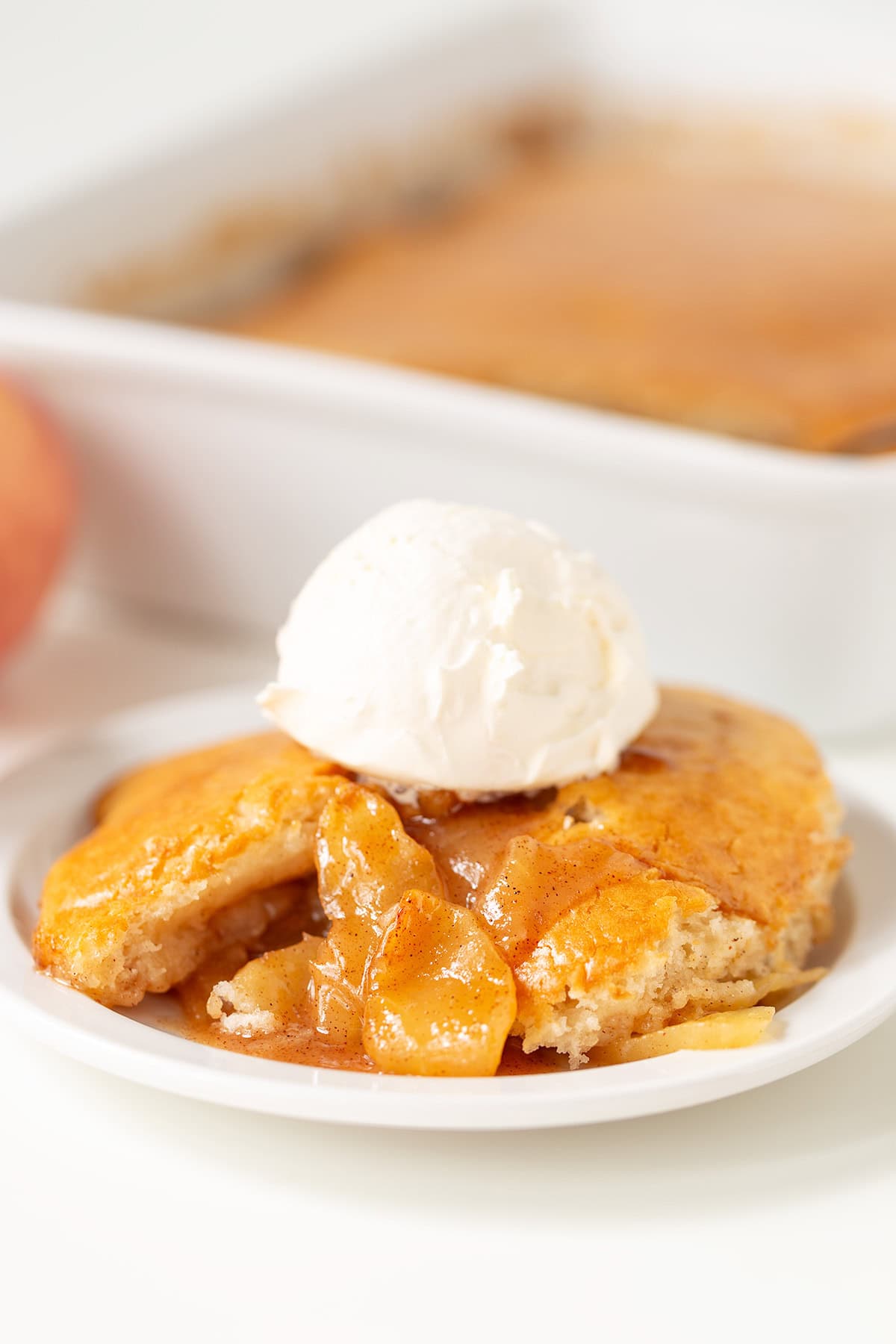 A serving of warm Apple Cobbler topped with a scoop of vanilla ice cream on a white plate, with the baking dish in the background.