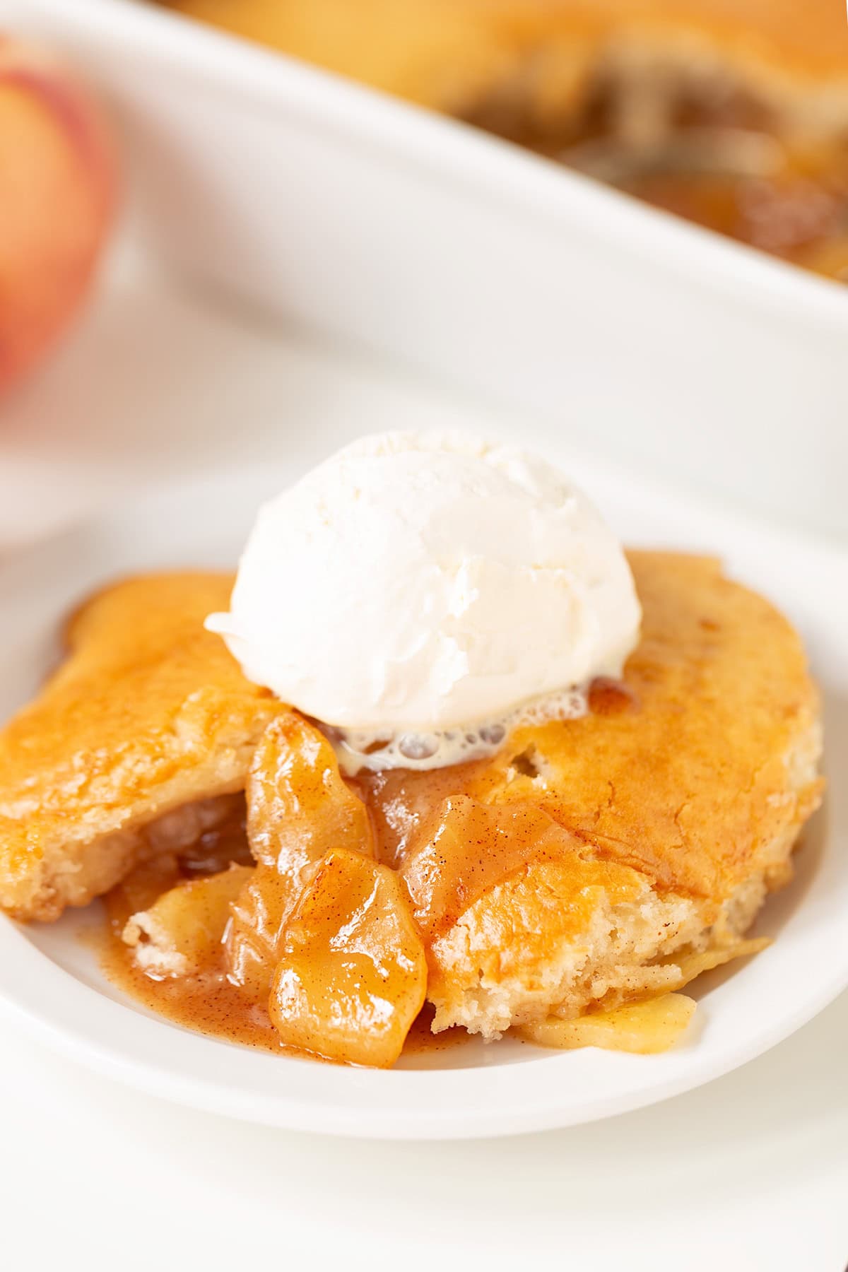 A serving of apple cobbler with visible apple slices, reminiscent of classic apple cobbler, topped with a scoop of vanilla ice cream on a white plate.