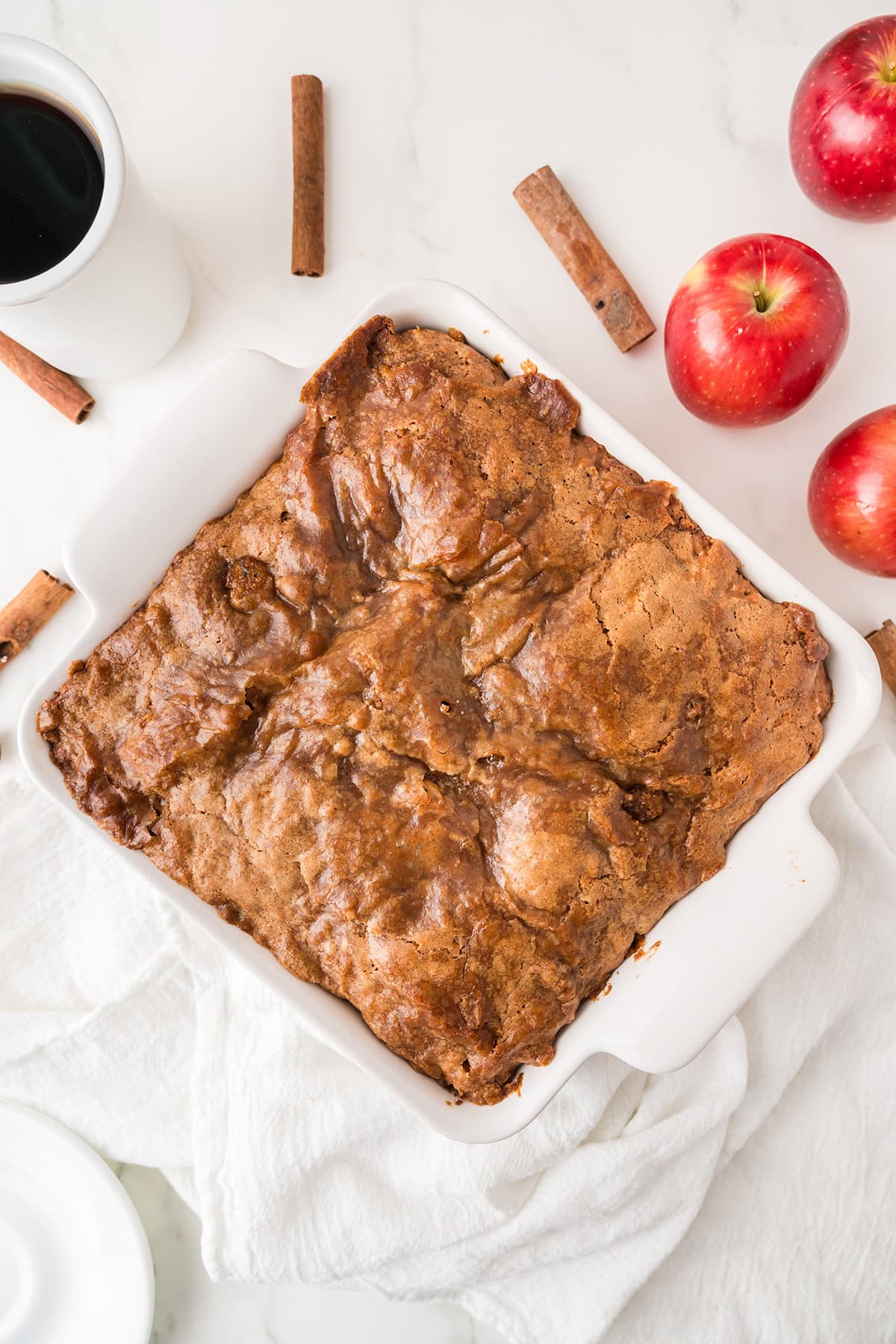 A baked apple coffee cake dessert in a white square dish sits on a white cloth, with three red apples, cinnamon sticks, and a cup of coffee on a marble surface nearby.
