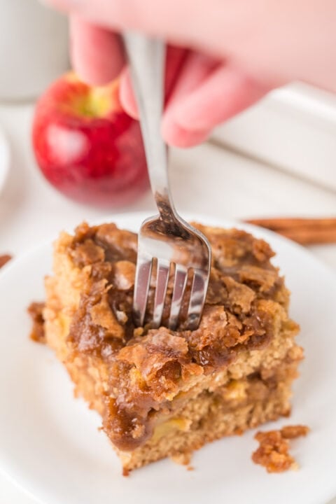 A hand holds a fork, cutting into a slice of crumb-topped Apple Coffee Cake on a white plate, with a red apple and cinnamon sticks in the background.