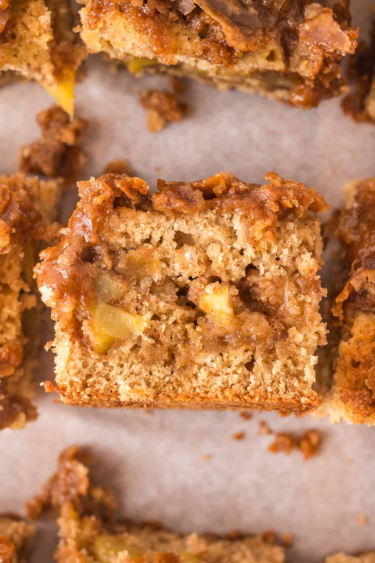 A close-up view of a square slice of moist Apple Coffee Cake with visible apple chunks and a crumbly brown topping, placed on parchment paper.