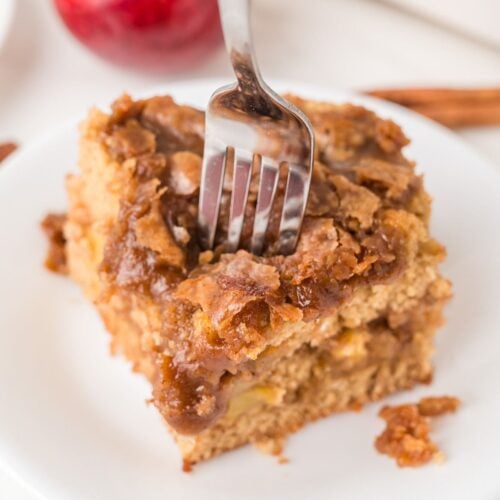 A fork is inserted into a square slice of apple coffee cake with a crumbly topping, served on a white plate.