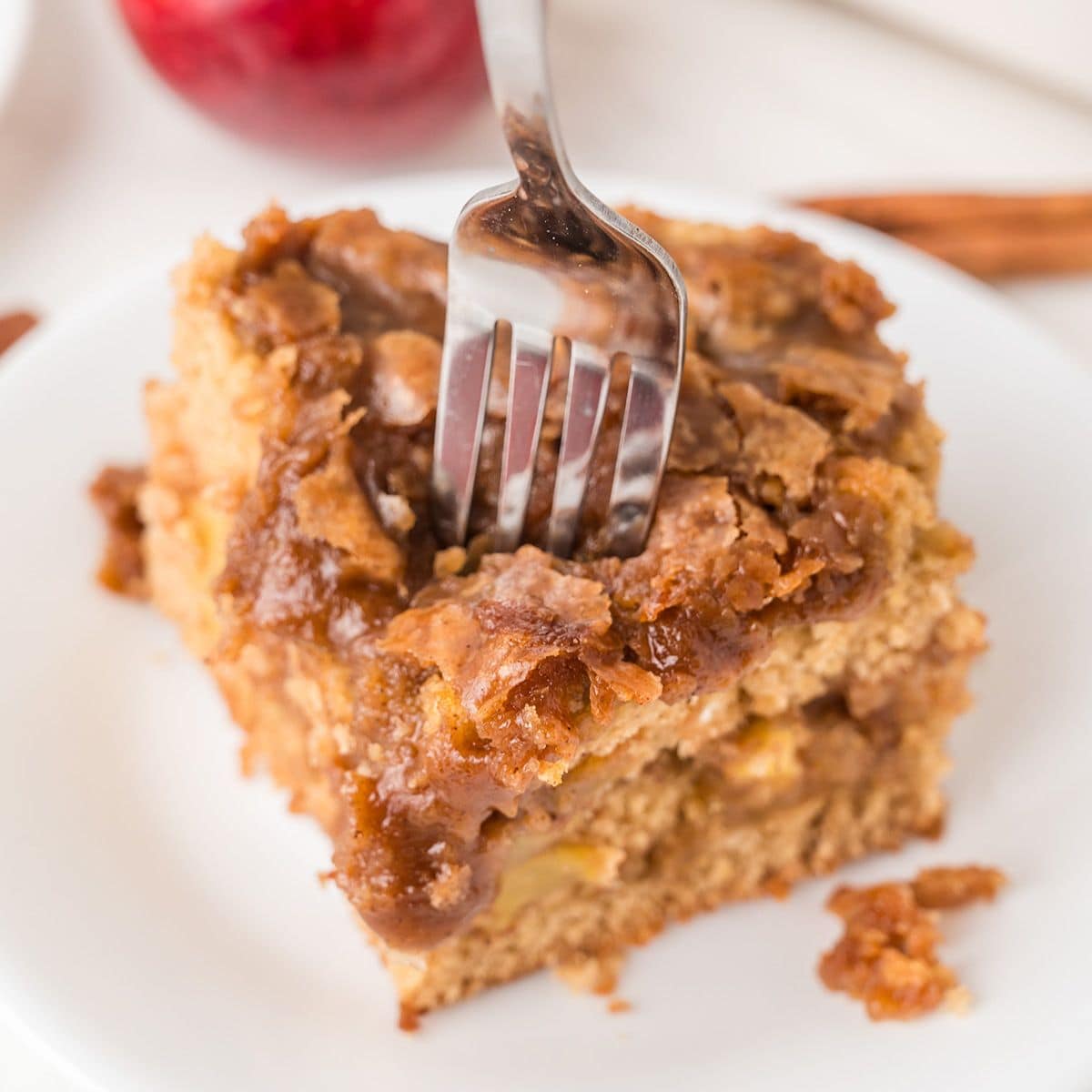 A fork is inserted into a square slice of apple coffee cake with a crumbly topping, served on a white plate.