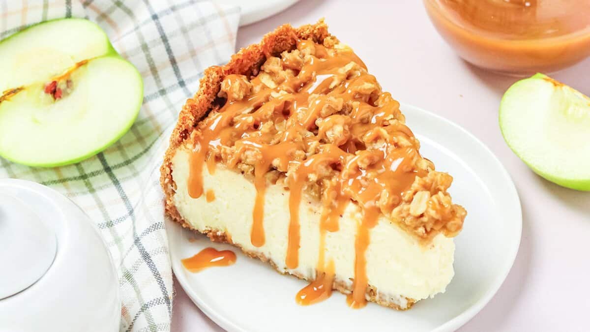 A slice of cheesecake with a crumbly topping and caramel drizzle on a white plate, next to green apple slices and a checkered cloth—perfect for those who crave Autumn desserts.