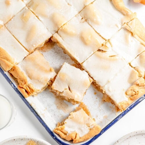 A baked pan of glazed apple bars, reminiscent of a classic Apple Slab Pie, cut into squares with a few pieces served on nearby plates, viewed from above.