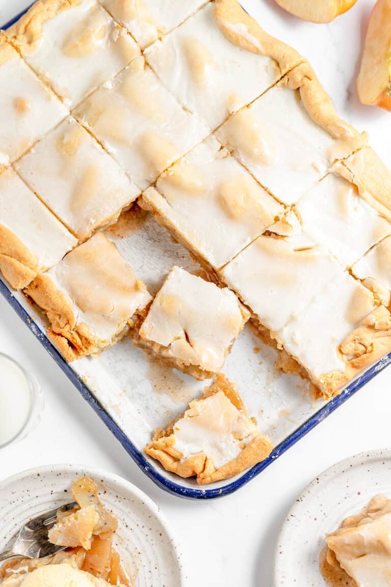 A baked pan of glazed apple bars, reminiscent of a classic Apple Slab Pie, cut into squares with a few pieces served on nearby plates, viewed from above.