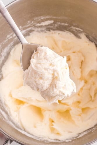 A metal spoon holds a scoop of creamy, whipped marshmallow frosting above a mixing bowl filled with more easy frosting.