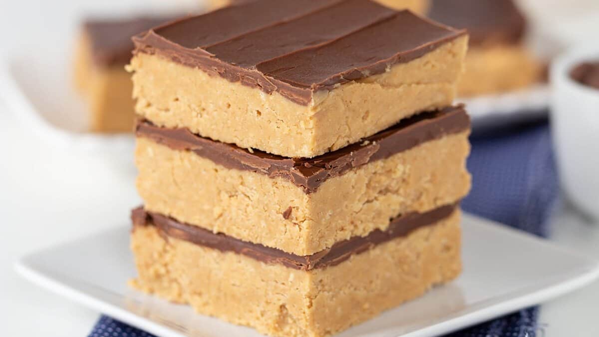 Three thick peanut butter bars with a chocolate layer on top are stacked on a white plate. Another plate with more bars is visible in the background, making these perfect seasonal sweets for your collection of Autumn desserts.